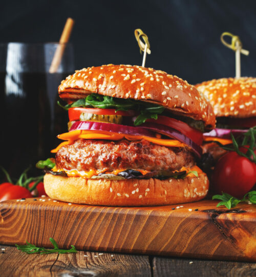 Two delicious homemade burgers of beef, cheese and vegetables on an old wooden table. Fat unhealthy food close-up.